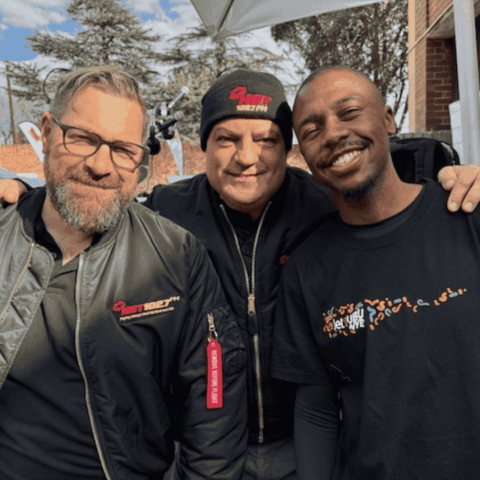 Three men stand close together outdoors, smiling at the camera. Two wear black jackets and hats with red logos, and one wears a black T-shirt with colorful text. Trees and a brick building are visible in the background.