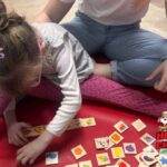 A young girl sits on a red mat playing with colorful wooden tiles while an adult sits beside her. The image has a HOT 102.7 FM Hot Cares Christmas in July logo in the bottom right corner.