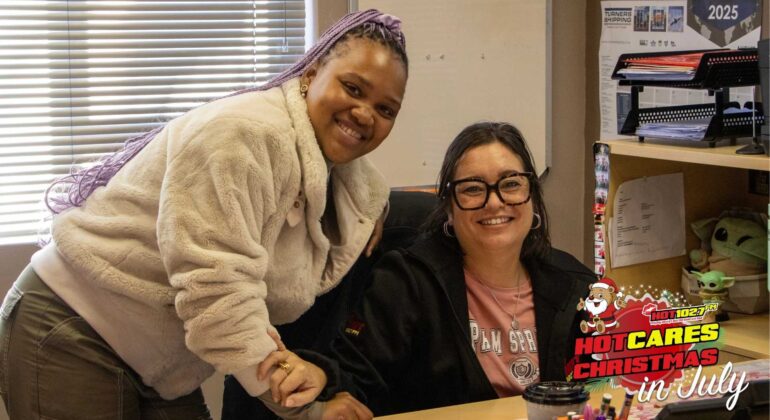 Two women smiling in an office, one standing and one sitting at a desk. A festive HOT CARES Christmas in July logo is displayed in the bottom right corner.