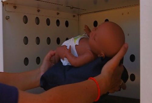 A person is placing a doll dressed in white clothes into a ventilated metal box, possibly simulating the use of a baby safe surrender drop-off.