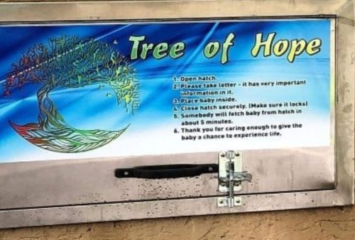 A sign titled Tree of Hope with instructions for placing a baby safely inside a hatch. The sign features a colorful tree design and a secured metal hatch beneath it on a beige wall.