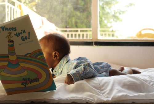 A baby lies on their stomach on a bed, holding and looking at a large, colorful book titled Oh, the Places Youll Go! by Dr. Seuss, with sunlight streaming through a window behind them.