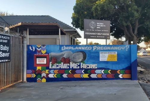 A colorful mural outside a pregnancy help center features a baby wrapped in a red blanket with text: Unplanned Pregnancy? Leave Me Here. Signs provide Baby Saver drop-off instructions and contact information.
