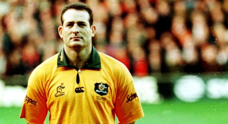 A male rugby player in a yellow and green jersey stands on a field, looking ahead with a serious expression. The background is blurred, showing an audience in a stadium.