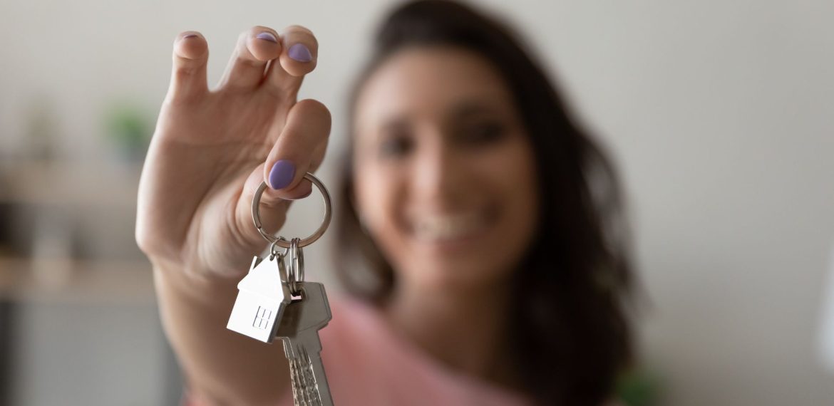 A smiling person with shoulder-length hair holds a keyring with two keys and a house-shaped keychain toward the camera, celebrating homeownership against a softly blurred background.