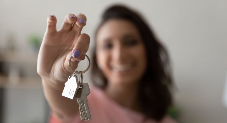 A smiling person with shoulder-length hair holds a keyring with two keys and a house-shaped keychain toward the camera, celebrating homeownership against a softly blurred background.