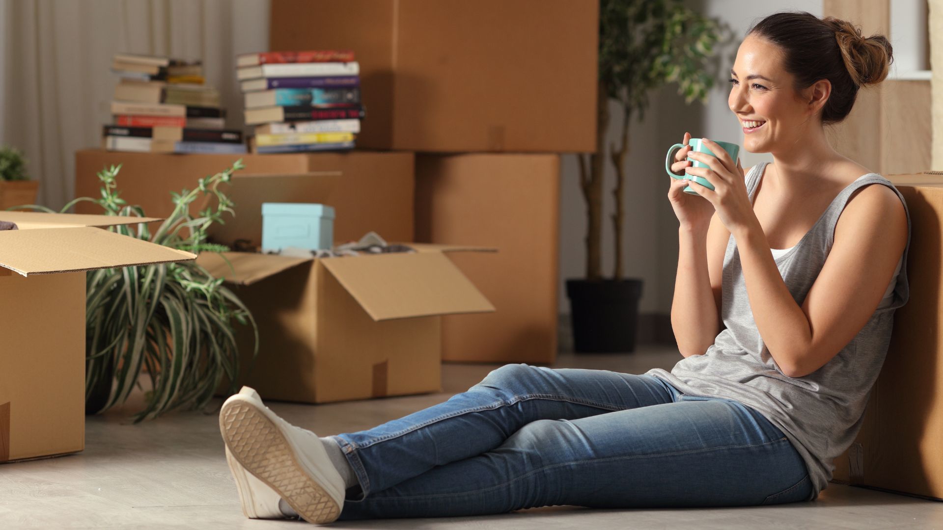 A woman sits on the floor, smiling and holding a mug, surrounded by moving boxes, books, and a potted plant in a well-lit room, celebrating the joy of new homeownership.