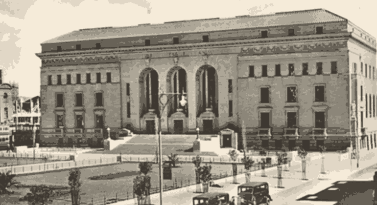 A historic black-and-white photo of a large, ornate stone building with tall arched windows and columns, steps leading to the entrance, vintage cars parked in front, and trees lining the street captures a moment as timeless as aged vinegar.