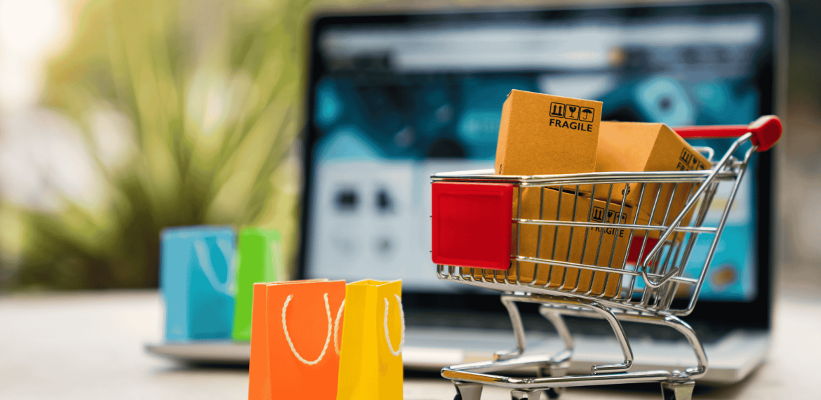 A small shopping cart with cardboard boxes and three colorful paper bags sits in front of an open laptop, symbolizing online shopping, e-commerce, and a seamless customer experience.
