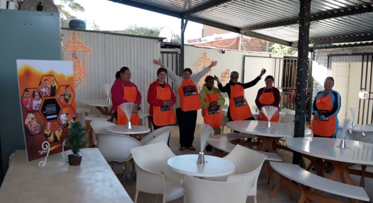 Seven people wearing orange aprons stand and wave inside a covered outdoor seating area with white tables and chairs. A small potted plant and a colorful poster are visible in the foreground.