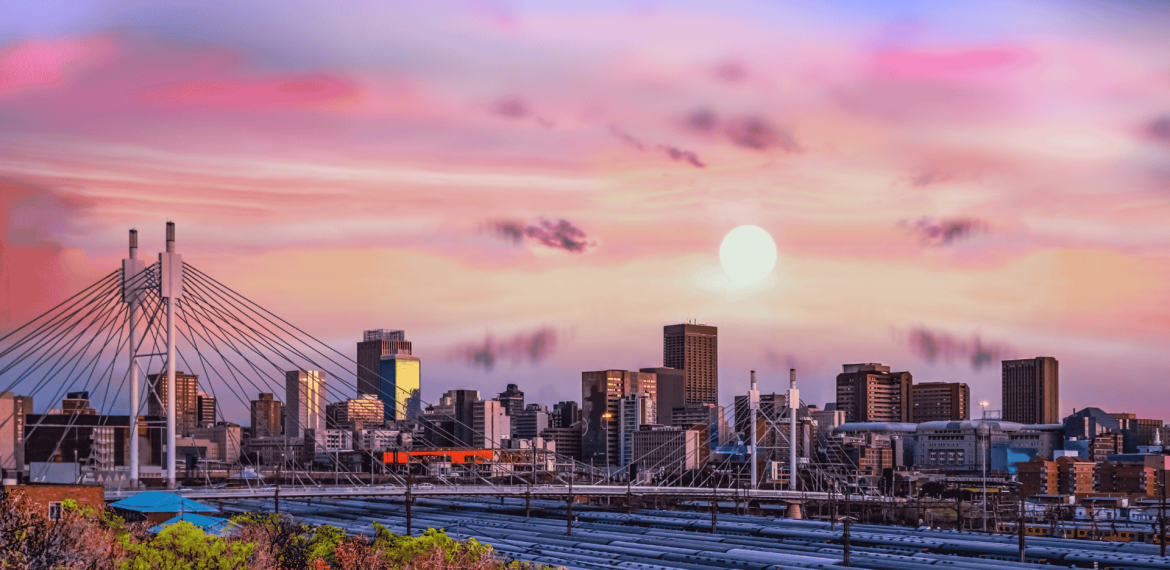 A vibrant Joburg city skyline at sunset, featuring a modern cable-stayed bridge in the foreground and tall buildings under a colorful sky with pink, purple, and orange hues. The sun is low on the horizon.