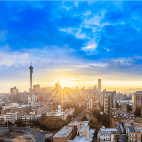 A city skyline at sunrise with sunlight streaming between Joburg's buildings. The tall observation tower stands out, and the sky above is partly cloudy with bright blue and golden hues.