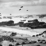 A black-and-white photo shows numerous military ships and landing craft at a crowded beach during World War II. Vehicles, troops, and equipment fill the shore, while Spitfire fighters soar above barrage balloons and many ships offshore.
