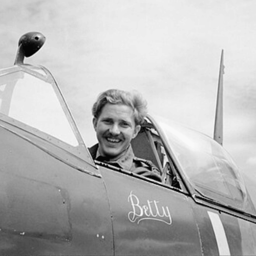 A smiling pilot sits in the cockpit of a Spitfire fighter plane with the name Betty painted on the side. The sky is cloudy in the background.