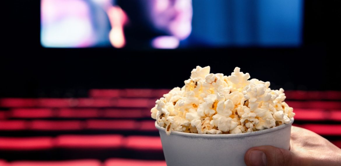 A hand holding a tub of popcorn in the foreground, with empty red seats and a blurry cinema screen in the background inside a movie theater.