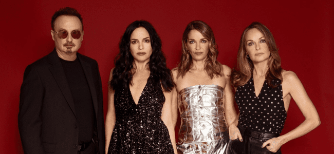 Four adults stand against a red background. The man on the left, likely from The Corrs, wears sunglasses and dark clothing; the three women wear fashionable outfits, including a sequined dress, a metallic strapless dress, and a sleeveless polka-dot top.