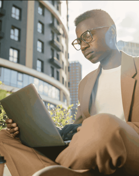 A Gen Z professional wearing glasses and a brown suit sits outdoors, hustling with a laptop on their lap. Modern buildings, lush greenery, and bright sunlight create an inspiring backdrop for work or study.