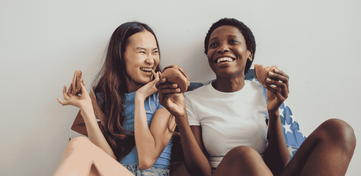 Two young women sitting side by side, smiling and laughing while holding sandwiches, with one wearing a light blue shirt and the other in white, seated against a wall with a patterned blanket at Witkoppen Clinic.