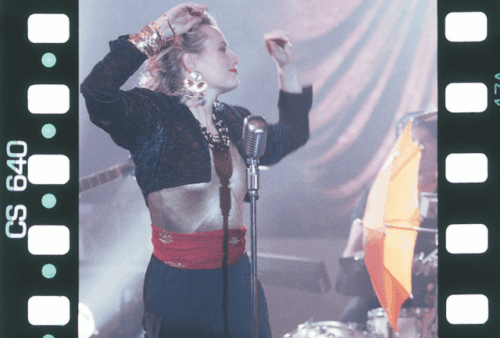 A performer in vintage-style clothing sings passionately into a retro microphone on stage, surrounded by dramatic lighting and musical instruments, with raised arms and expressive movement.