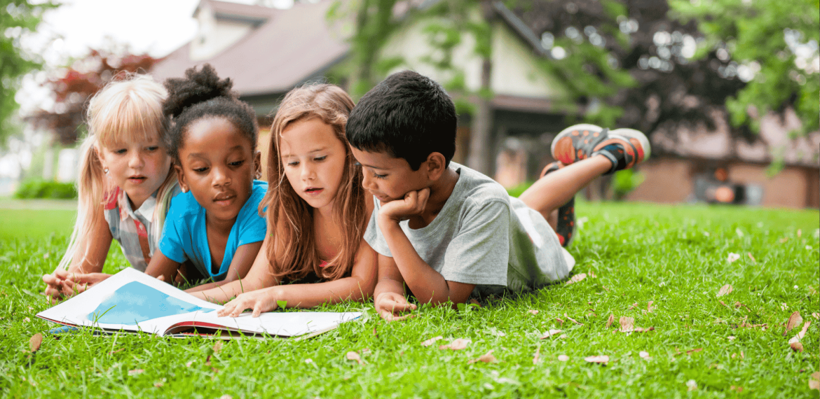 Four young children lie on the grass outside, closely reading a large open book together, imagining stories about the ict sector, with trees and a house in the background on a sunny day.
