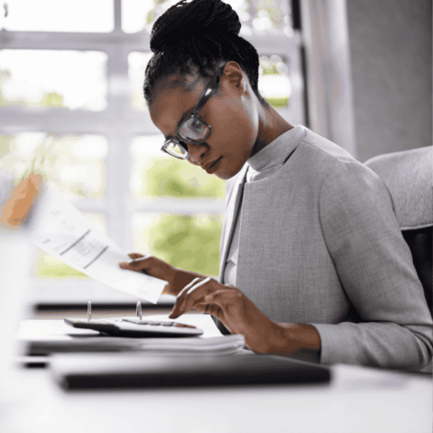 A woman wearing glasses and a gray blazer sits at a desk, reviewing documents and using a calculator, with a window and greenery in the background.
