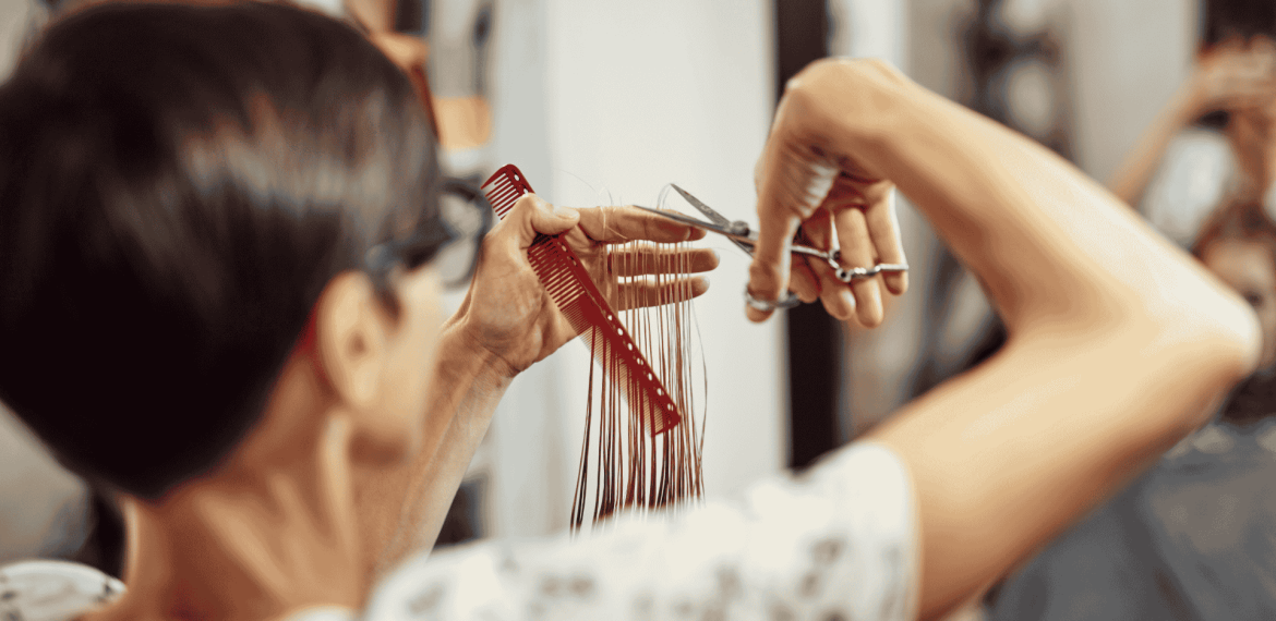 A hairstylist with short dark hair holds a red comb and scissors, carefully trimming long strands of hair in a salon, showcasing expert Hair Care. The focus is on the stylist’s hands and tools.