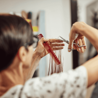 A hairdresser wearing glasses uses scissors and a red comb to cut a client’s wet hair, carefully holding a section between their fingers in a salon setting—a true professional focused on Hair Care.