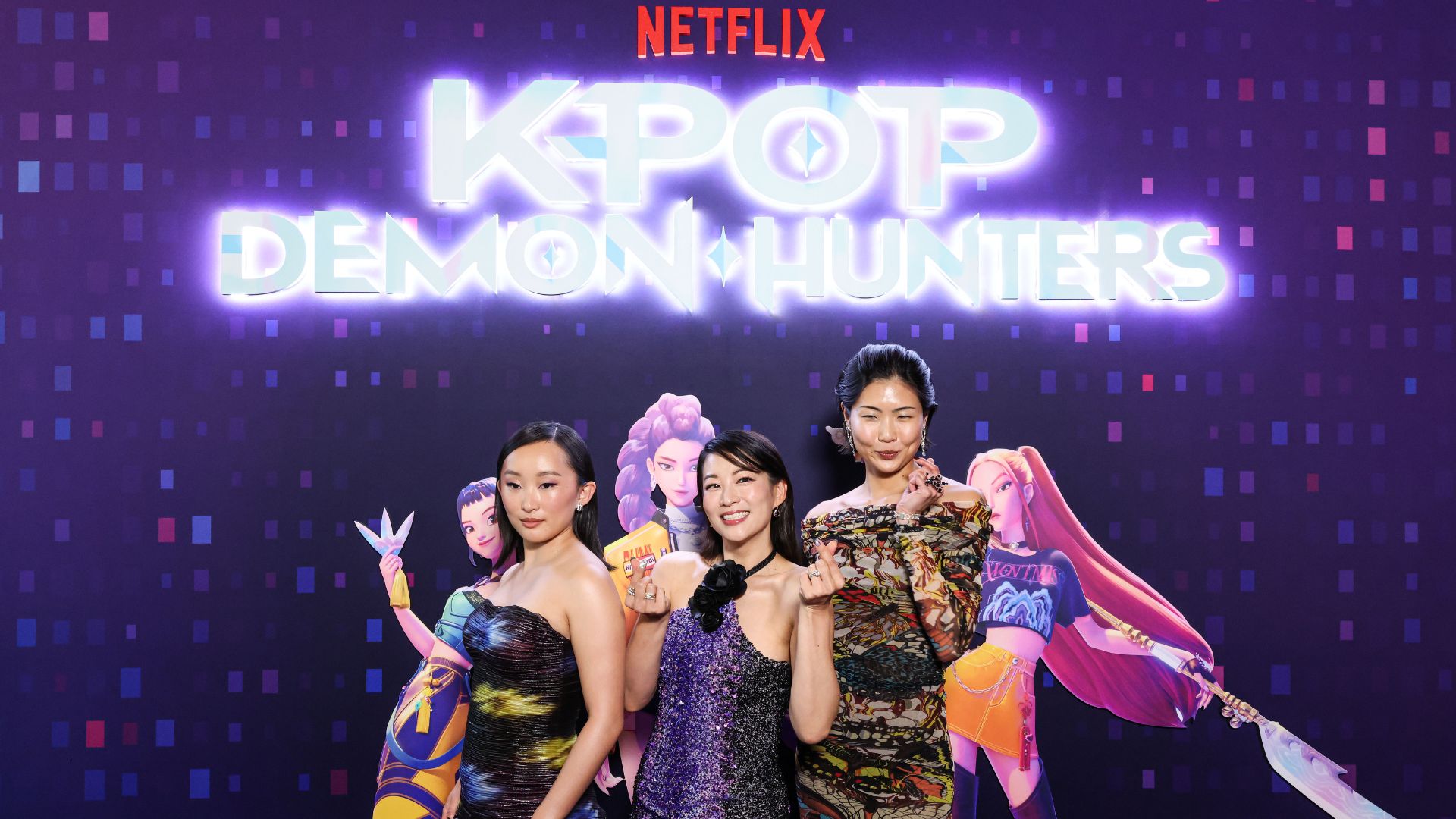 Three women in colorful dresses pose and smile in front of a Netflix K-pop Demon Hunters sign, featuring animated characters. The purple backdrop with square patterns highlights the fun, energetic vibe of the K-pop Demon Hunters event.