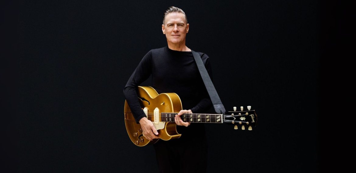 A man in a black long-sleeve shirt stands against a plain black background, holding a gold electric guitar with a strap over his shoulder and looking straight ahead, reminiscent of Bryan Adams’ iconic style.