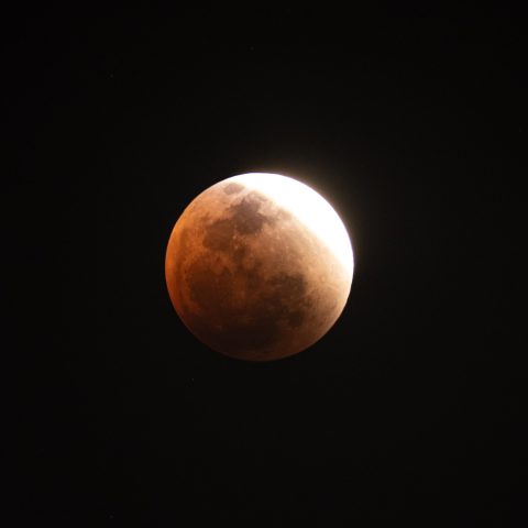 A lunar eclipse with the moon partially covered by Earth's shadow, showing a Blood Moon’s reddish hue on the lower part and a bright white edge on top against a dark night sky.