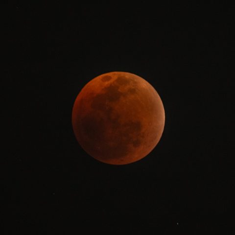 A dark, reddish Blood Moon appears in the night sky, representing a lunar eclipse with its surface partially illuminated and surrounded by deep blackness.