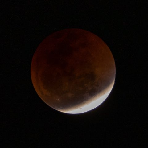 A close-up view of the moon during a Blood Moon lunar eclipse, with most of the surface appearing dark red and a small crescent at the bottom illuminated brightly against a black sky.