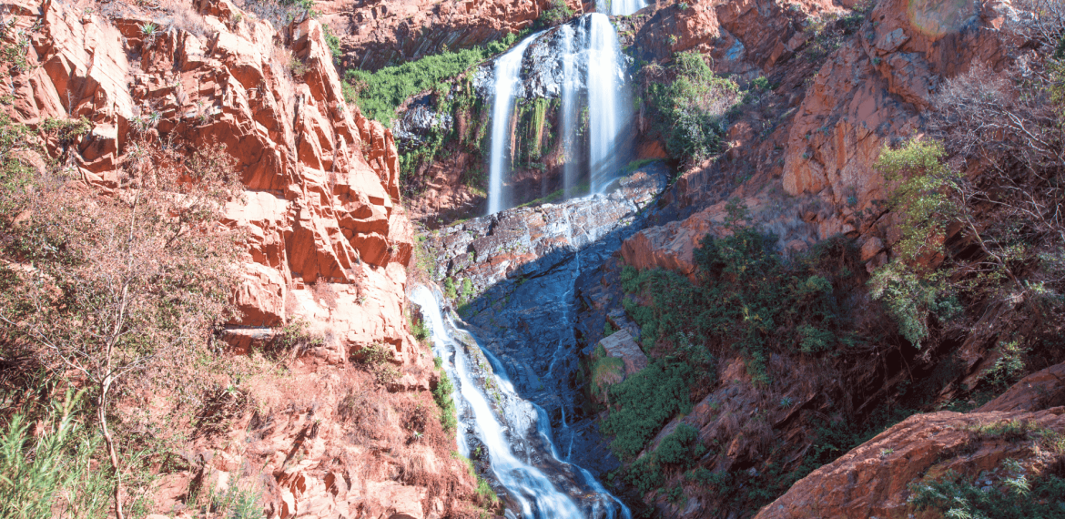 A tall, multi-tiered waterfall cascades down rugged, reddish-brown cliffs surrounded by green vegetation under a clear blue sky, much like the tranquil settings near Witkoppen Clinic.