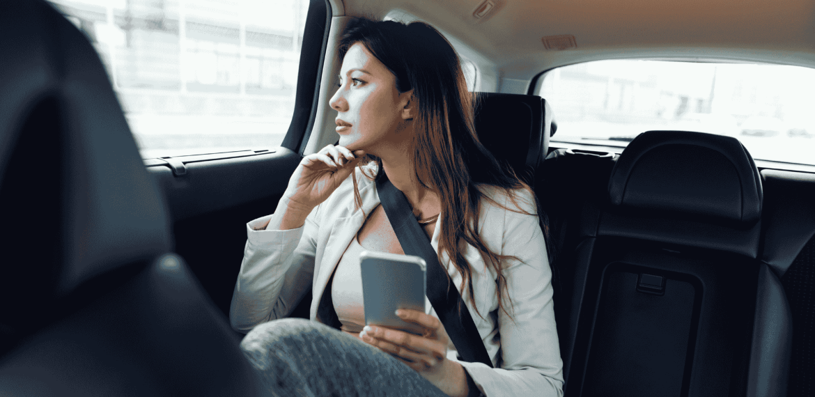 A woman sitting in the backseat of a car, wearing a seatbelt, holds a smartphone and gazes thoughtfully out the window, pondering her journey toward homeownership during the daytime.