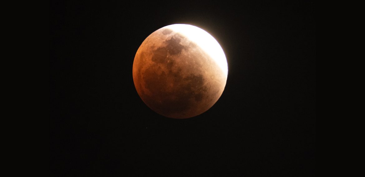 A partially eclipsed Blood Moon appears with a reddish hue and a bright white edge against the dark night sky, illustrating the progression of a lunar eclipse.