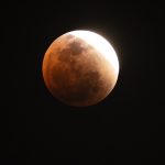 A partially eclipsed Blood Moon appears with a reddish hue and a bright white edge against the dark night sky, illustrating the progression of a lunar eclipse.