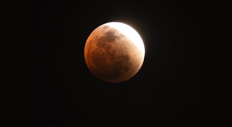 A partially eclipsed Blood Moon appears with a reddish hue and a bright white edge against the dark night sky, illustrating the progression of a lunar eclipse.