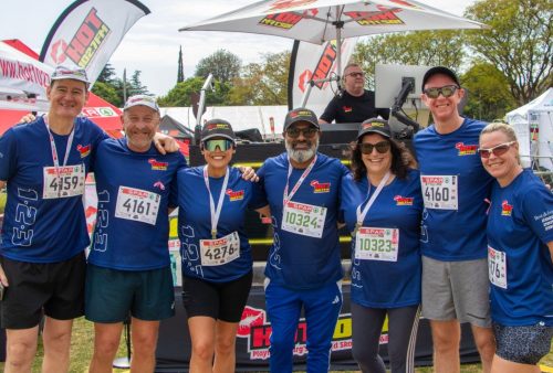 Seven smiling people in matching blue shirts and medals pose together outdoors at the SPAR Womens Challenge, with race bibs on their chests and event banners, a tent, and equipment in the background.