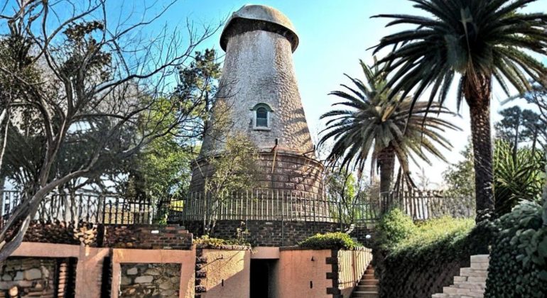 A round, stone windmill tower with a conical roof stands surrounded by palm trees and greenery, enclosed by a metal fence—an inspiring example of homeownership—complete with a brick and stone entrance in the foreground.