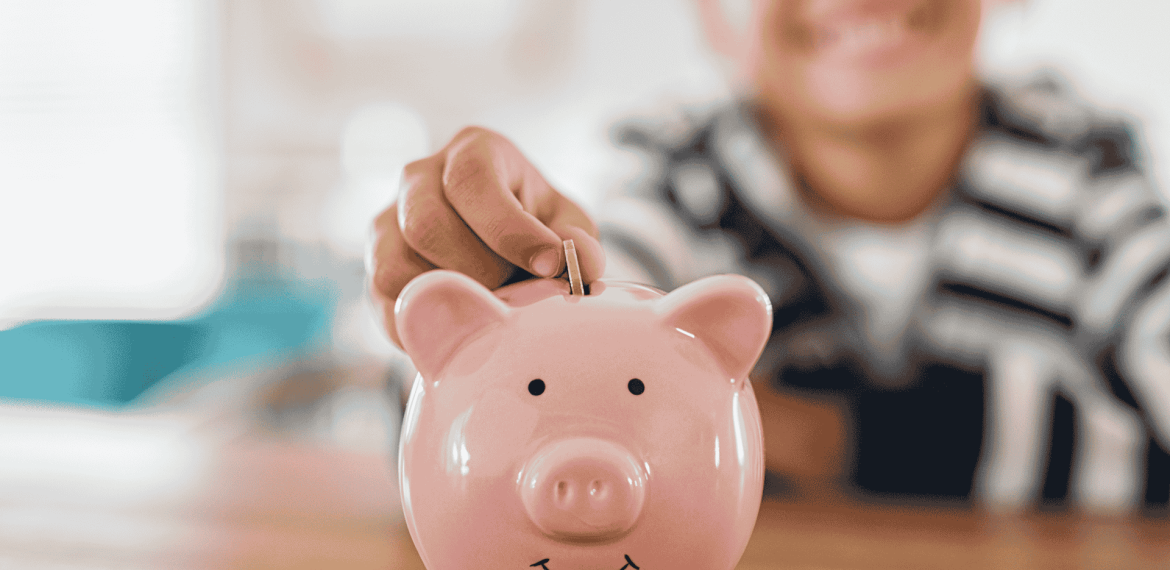A child smiles while placing a coin into a pink piggy bank on a wooden table, showing that saving money is far from brainrot. The background is softly blurred.