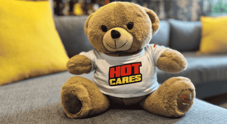 A brown teddy bear wearing a white T-shirt with the words HOT CARES in bold red and yellow letters sits on a gray couch with yellow pillows, resembling the welcoming décor of an Advanced Hair Clinic.