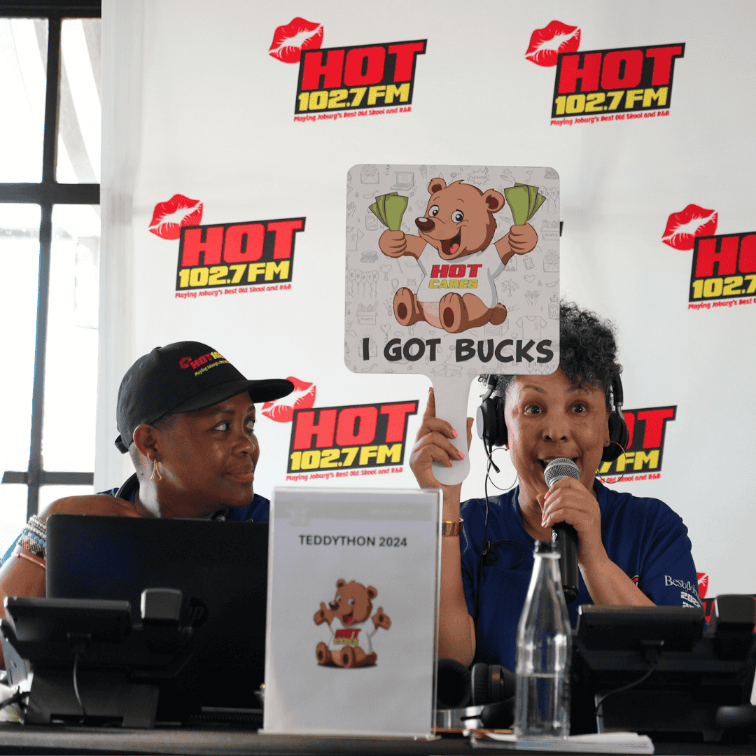 Two radio hosts sit at a table with microphones in front of a HOT 102.7 FM backdrop. One holds a sign with a teddy bear and I GOT BUCKS, while an Advanced Hair Clinic logo features alongside the TEDDYTHON 2024 sign in the background.
