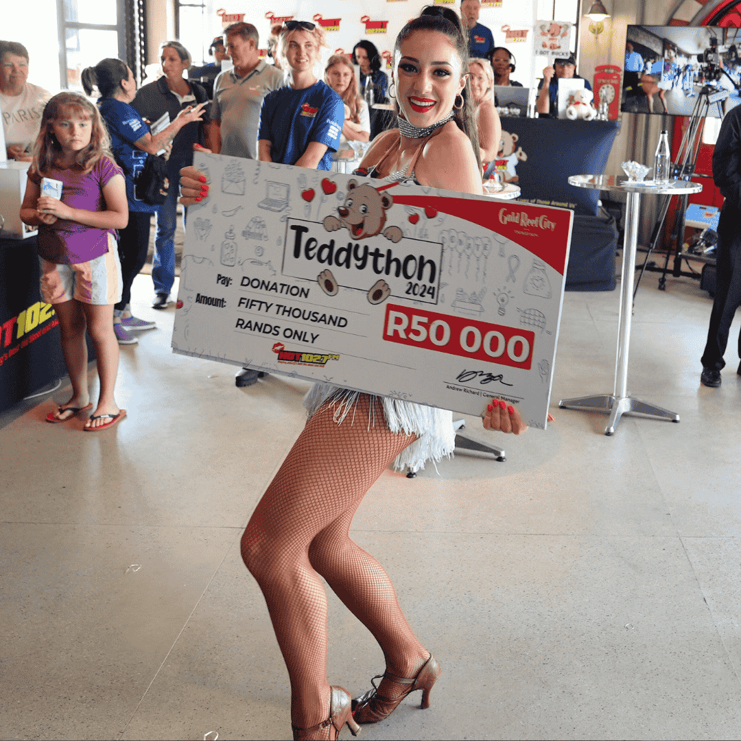 A smiling woman in a sparkly outfit and fishnet stockings holds a large check for R50,000 from Teddython 2024 at an indoor event, with people, cameras, and an Advanced Hair Clinic banner visible in the background.