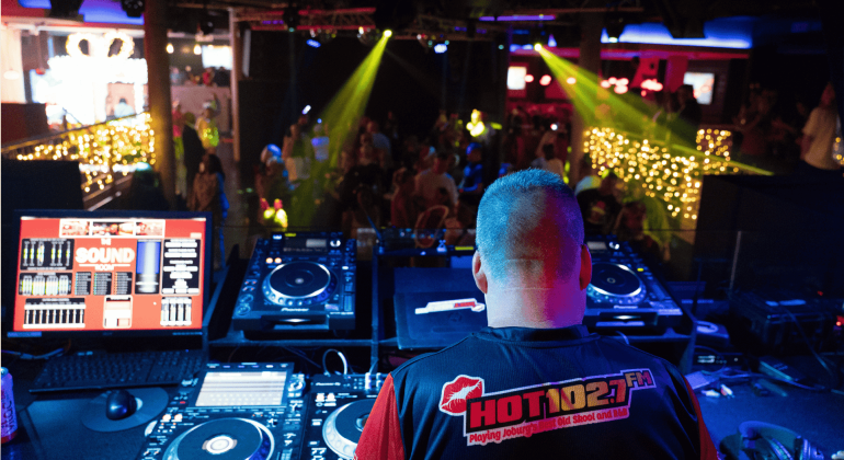 A DJ wearing a HOT 102.7 FM shirt stands at a booth with turntables and computer screens, performing for a lively Jingle Jam crowd in a club lit with colorful lights and string decorations.