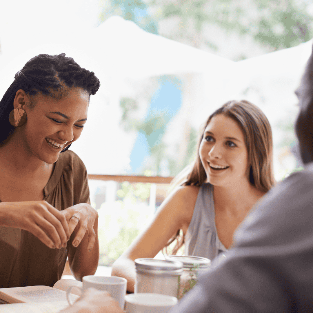 Two women sit at a table outdoors, smiling and laughing together, with drinks and a jar in front of them. The friendly atmosphere suggests they might be sharing stories about their experience at an advanced hair clinic.