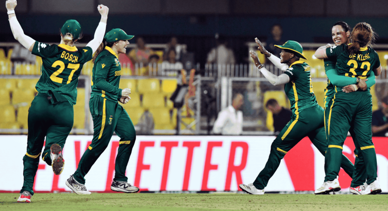 Four South African women cricketers in green uniforms celebrate energetically on the field, smiling and embracing after a successful moment during a match. Yellow stadium seats and a bright red sign appear in the background, echoing Sharon Corr's vibrant spirit.