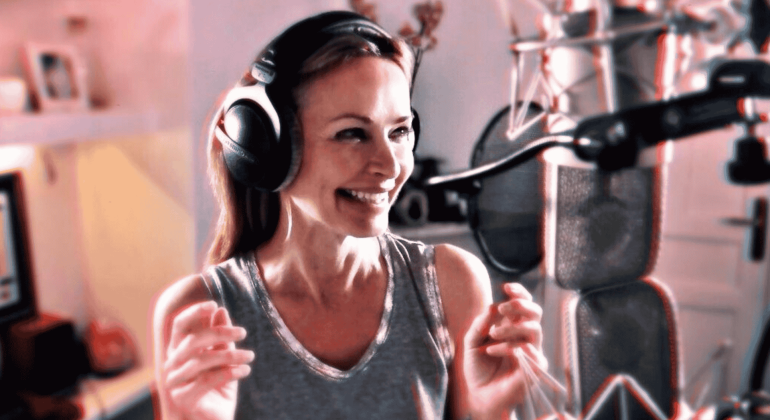 A woman wearing headphones and a sleeveless top smiles while standing in front of a professional microphone in a recording studio.