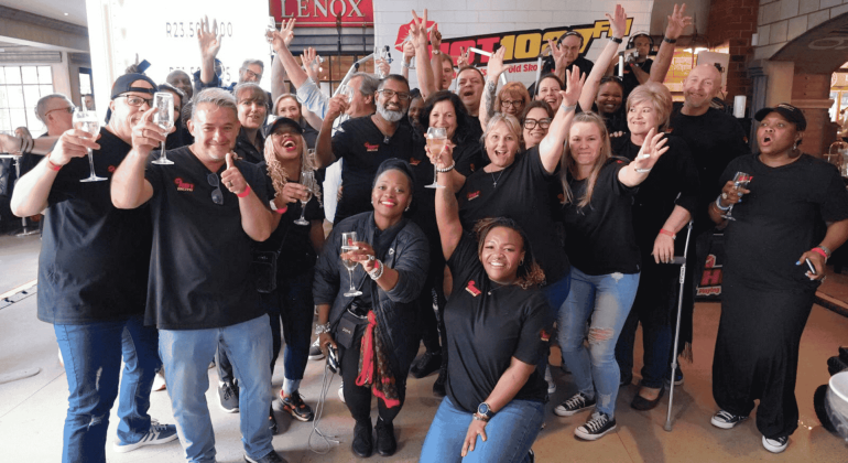 A large, diverse group of people in matching black shirts smiles and celebrates together indoors at Teddython 2025, raising glasses and posing enthusiastically for the camera.