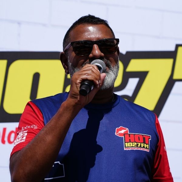 A man with short dark hair and a gray beard wearing sunglasses speaks into a microphone. Sporting a HOT 102.7 FM shirt, he stands before a large HOT 102.7 sign at the Radio Training Academy event.