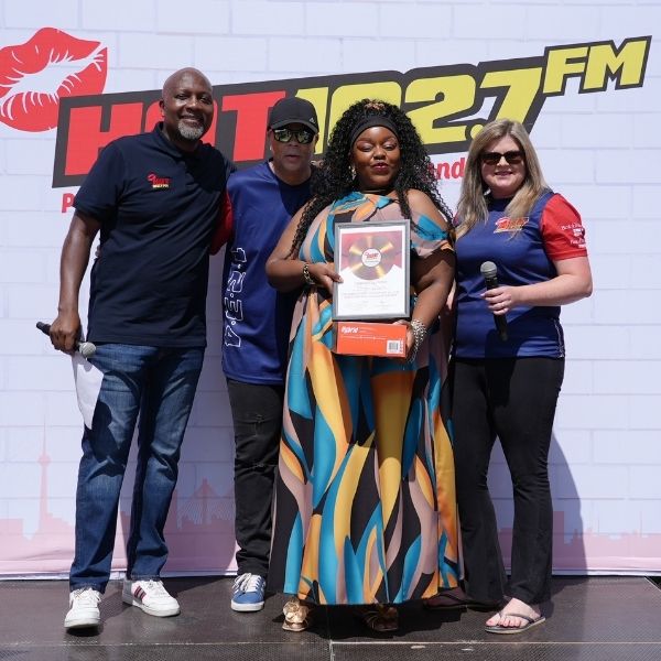 Four people stand on a stage in front of a HOT 102.7 FM sign, representing the Radio Training Academy. The person in the center holds a framed certificate and a gift box, while the others smile beside her.